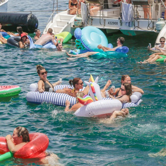 People relax on colorful inflatable floats, including a unicorn and a watermelon, in the water near anchored boats under sunny skies. Some hold drinks, and the atmosphere is festive and lively.