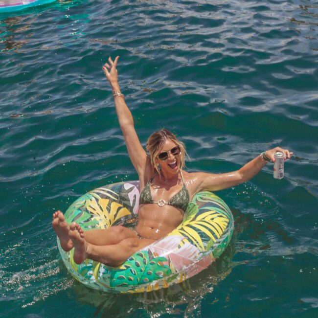 A woman in sunglasses and a bikini floats on a colorful inflatable ring in the water, smiling with one arm raised and holding a drink in the other hand. The sun is shining and there are more inflatables in the background.