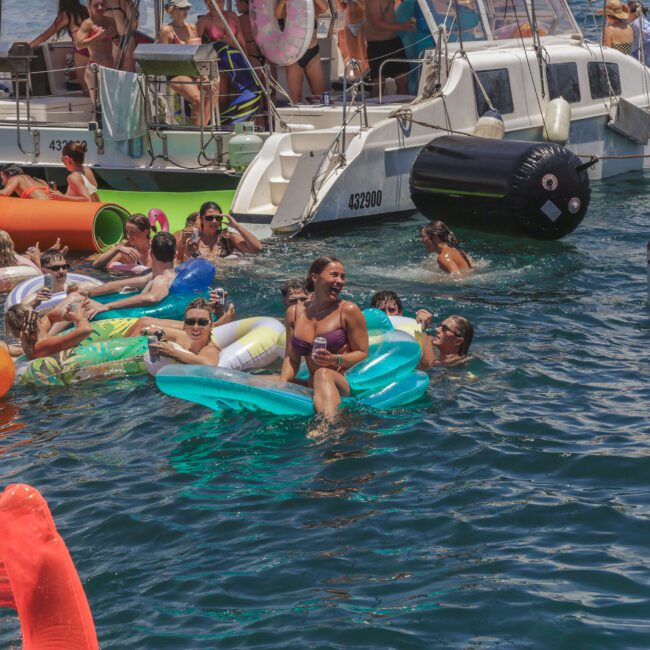 A group of people enjoy a pool party in the ocean, relaxing on colorful inflatables and swimming near anchored boats under sunny weather. The mood is festive and lively.