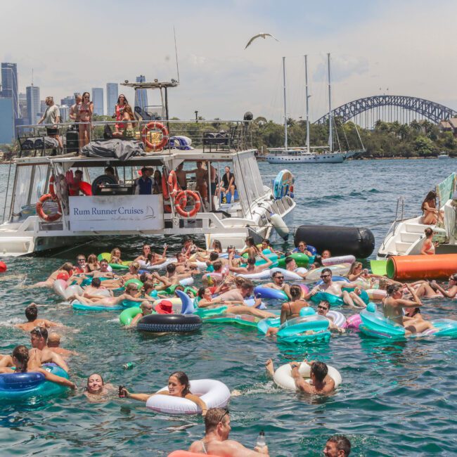 A large group of people on colorful inflatables float in the water near boats with "Rum Runner Cruises" signage, with a city skyline and Sydney Harbour Bridge visible in the background.