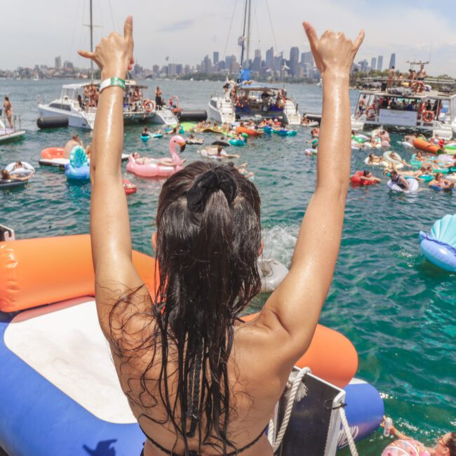 A woman with wet hair raises her hands in a "rock on" gesture at a lively boat party, surrounded by people on colorful inflatables in the water, with a city skyline visible in the background.
