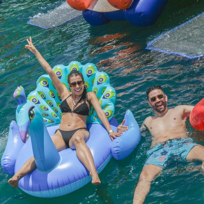 Two people smile and relax on colorful inflatable floats in the water; one woman on a blue peacock float raising a peace sign, and a man beside her on a red float, both enjoying a sunny day.