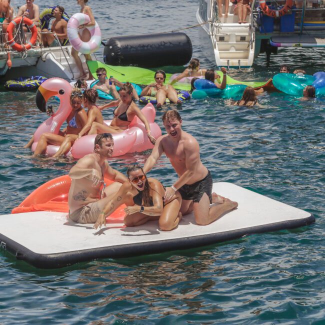 A group of people enjoying a sunny day on the water, with three people posing on a large floating mat surrounded by others relaxing on colorful inflatable rafts, boats, and pool floats in the background.