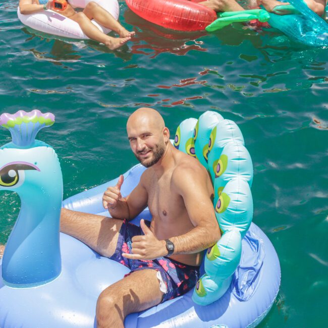 A man smiles and gives two thumbs up while sitting on a blue peacock-shaped pool float in clear green water, surrounded by other people on colorful floats. The logo "Yacht Social Club" is visible in the corner.
