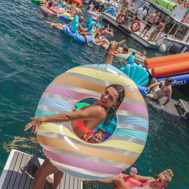 A woman in a colorful striped pool float smiles at the camera on a dock, with many people on inflatables and boats in the water behind her during a lively summer party.