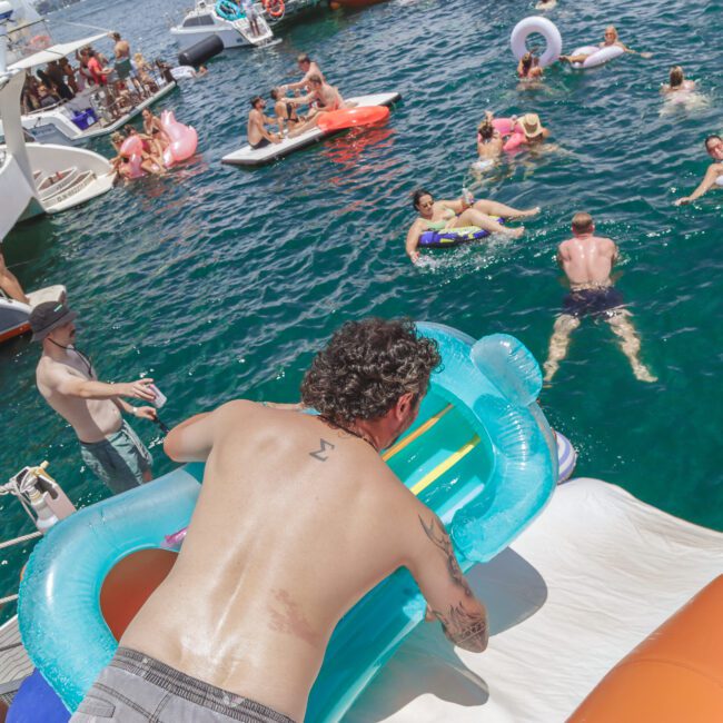 People enjoy a lively pool party near several boats, floating on inflatables and swimming in clear blue water. A man with a tattoo prepares to slide into the sea using an inflatable mattress.