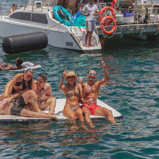 A group of five people relax and pose for the camera on a floating mat in the ocean near boats. Some are sitting and some are lying down, smiling and enjoying the sunny day on the water.