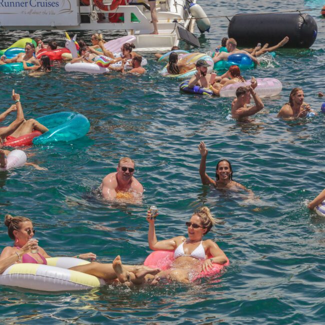 People relaxing and socializing on colorful pool floats in clear blue water near boats on a sunny day; many are holding drinks and enjoying the festive atmosphere.