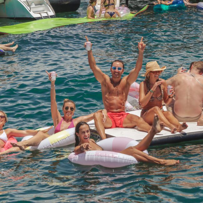 A group of smiling people in swimsuits relax on floats and a paddleboard in a busy, sunlit body of water, holding drinks and raising their arms in celebration among others enjoying a lively summer party.