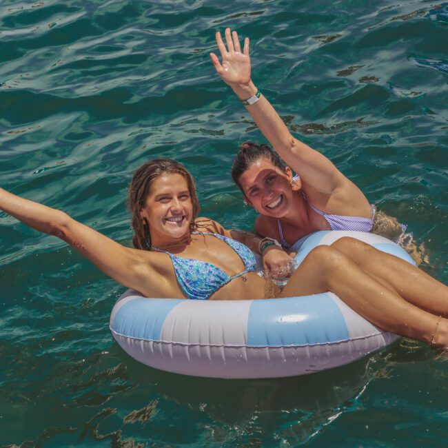 Two women in bikinis smile and wave while sitting together on a striped inflatable ring in clear blue water, enjoying a sunny day.