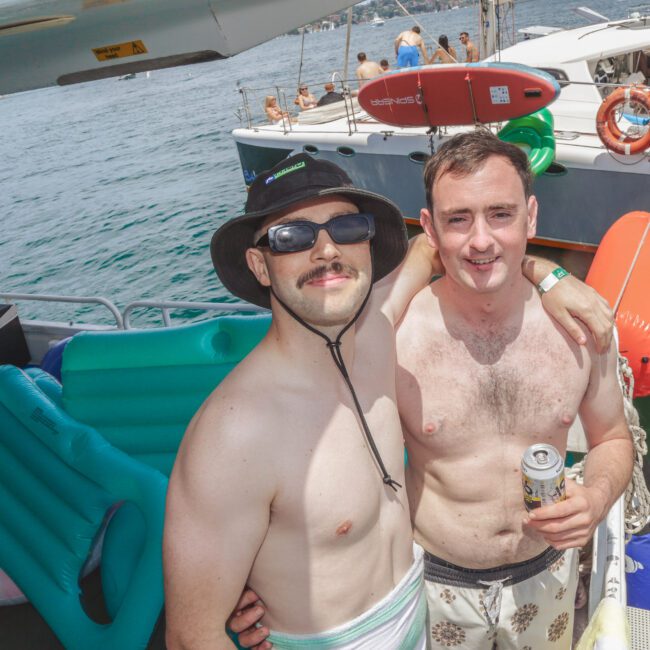Two men in swim trunks pose and smile on a boat, surrounded by inflatable pool floats. One wears a hat and sunglasses, and the other holds a drink. Other people and boats are visible on the water in the background.