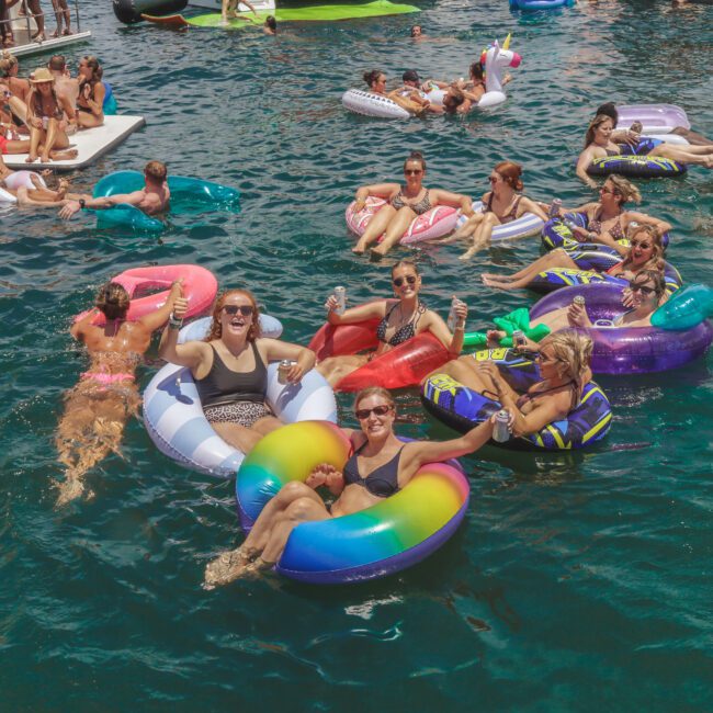 A group of people relax and smile on colorful inflatable floats in the water, surrounded by others on various floaties and boats, enjoying a sunny day. The scene is festive and lively.
