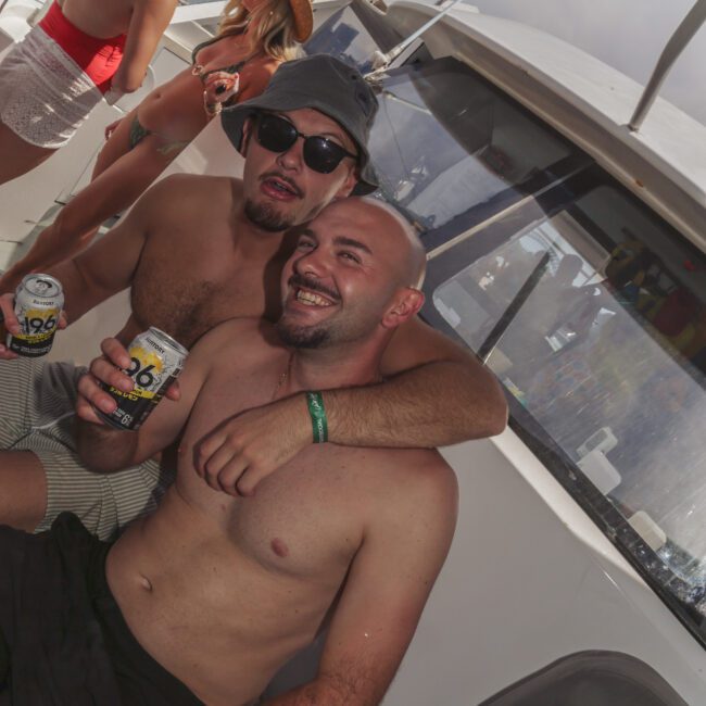 Two shirtless men sit closely on a boat, smiling and holding canned drinks. Other people are in the background, some standing and talking, with sunlight and water visible outside. The scene is lively and festive.