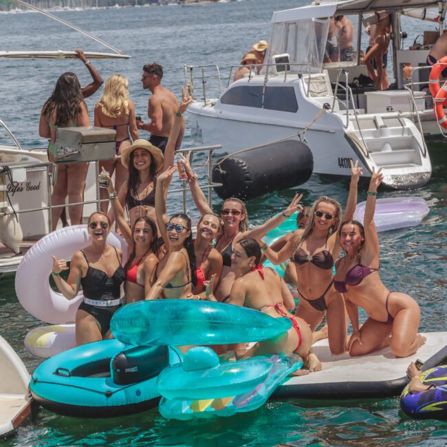 A group of people in swimsuits pose and smile on a floating platform with inflatable pool floats near docked boats on a sunny day. Others relax and socialize on nearby yachts in the background.