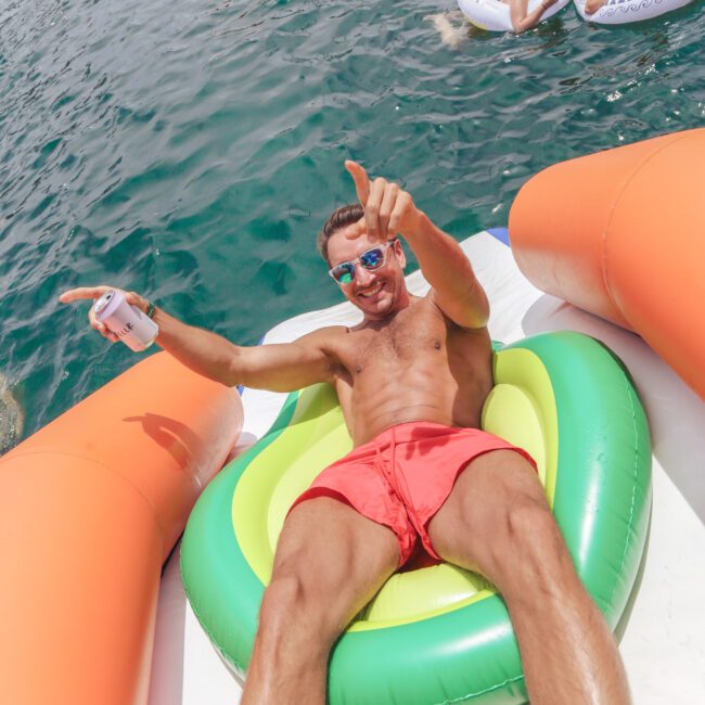 A man in red swim trunks lounges on an inflatable float in the water, holding a drink and pointing at the camera. Other people float nearby on colorful inflatables. The scene is sunny and festive.