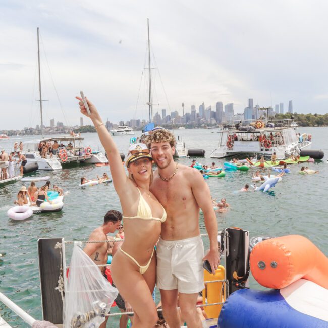 Two people in swimwear smile and pose on a boat at a lively floating party, with many people on boats and inflatables in the water and a city skyline visible in the background.
