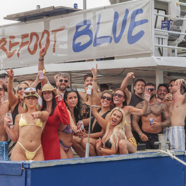 A group of people in swimwear happily pose and cheer on the deck of a boat labeled “Barefoot Blue.” Many are holding drinks, smiling, and making celebratory gestures, enjoying a lively outdoor party.