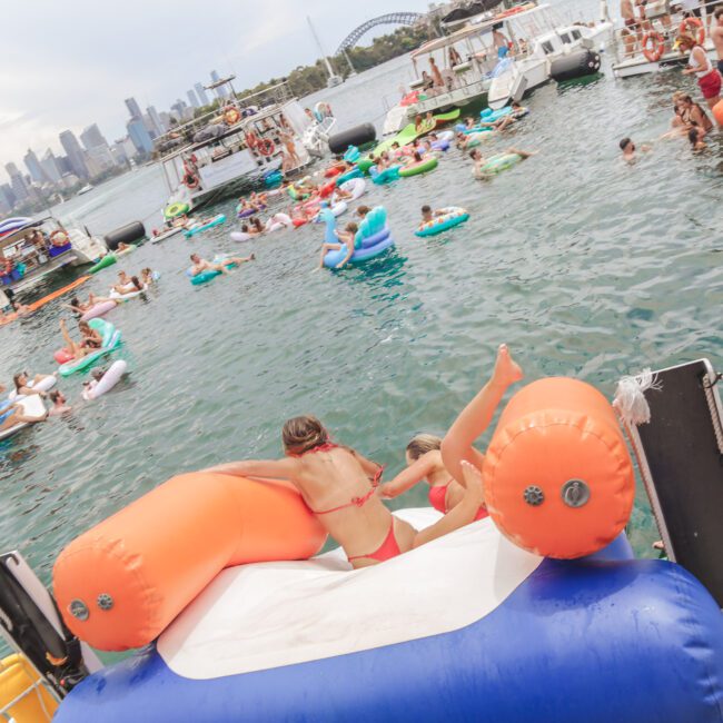 A crowded harbor scene with people relaxing on boats, inflatables, and floating devices in the water. The Sydney Harbour Bridge and city skyline are visible in the background under a cloudy sky.