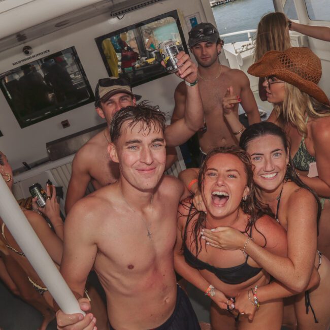 A group of young adults in swimwear smile and pose for a photo at a lively boat party, holding drinks and appearing joyful. Some people are dancing, while others look at the camera. Water and a dock are visible outside.