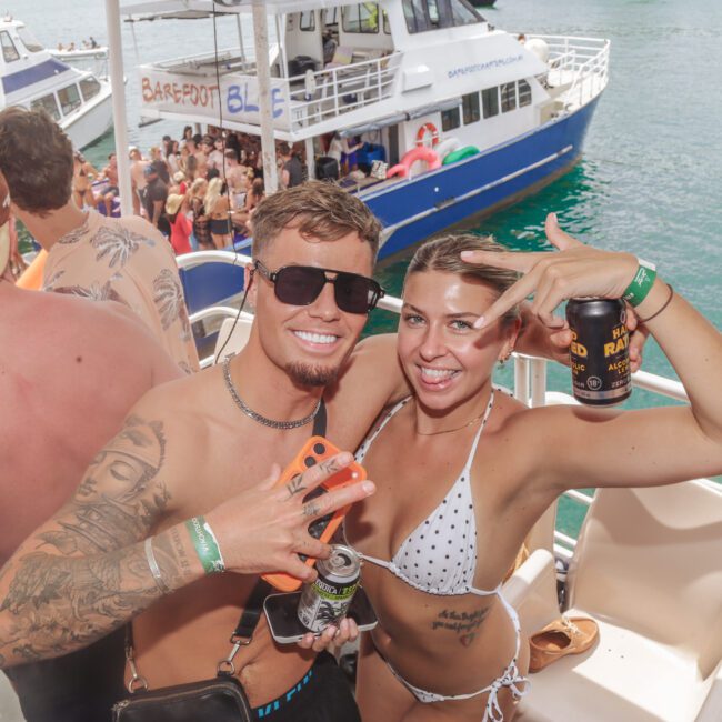 A smiling man and woman in swimsuits pose for the camera on a crowded party boat, holding drinks and making peace signs, with more people and boats in the background on the water.
