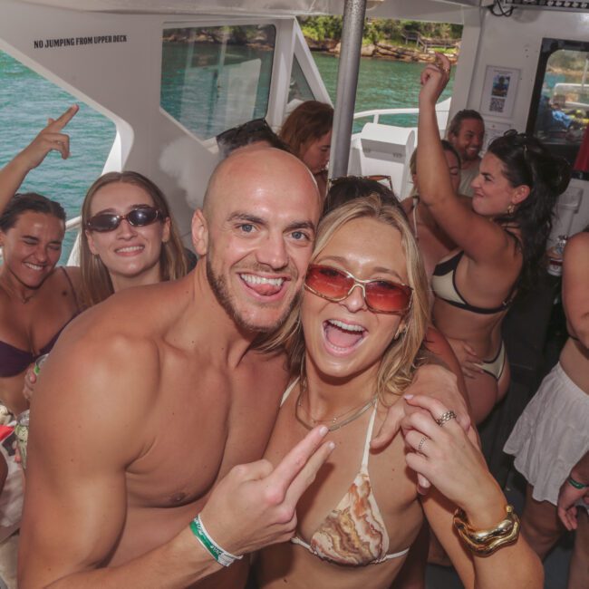 A group of people in swimsuits dance and smile on a boat, enjoying a sunny day on the water. Some make peace signs and hold drinks, while the background shows blue water and greenery outside the boat.
