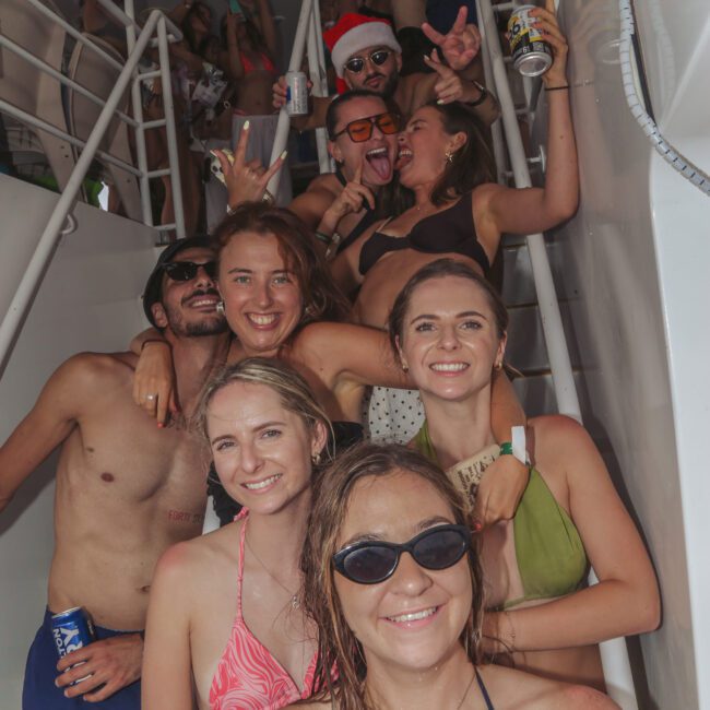 A group of smiling friends in swimsuits pose on a boat staircase during a lively party. Some people in the background hold drinks, and one person is wearing a Santa hat. The mood is festive and fun.