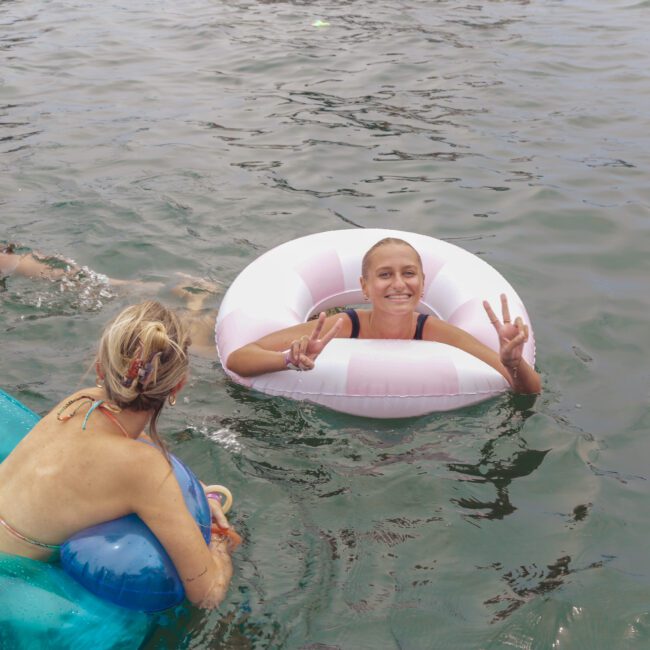 Three women enjoy swimming in a lake; one is floating in a white inner tube and flashing a peace sign, another swims nearby, and the third sits in a blue float. The water is calm, and all are smiling.