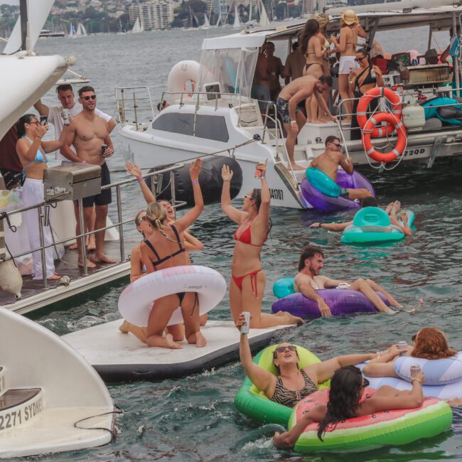 A group of people in swimsuits relax and have fun on colorful inflatables and boats in the water at a lively boat party, with music, drinks, and a festive atmosphere.