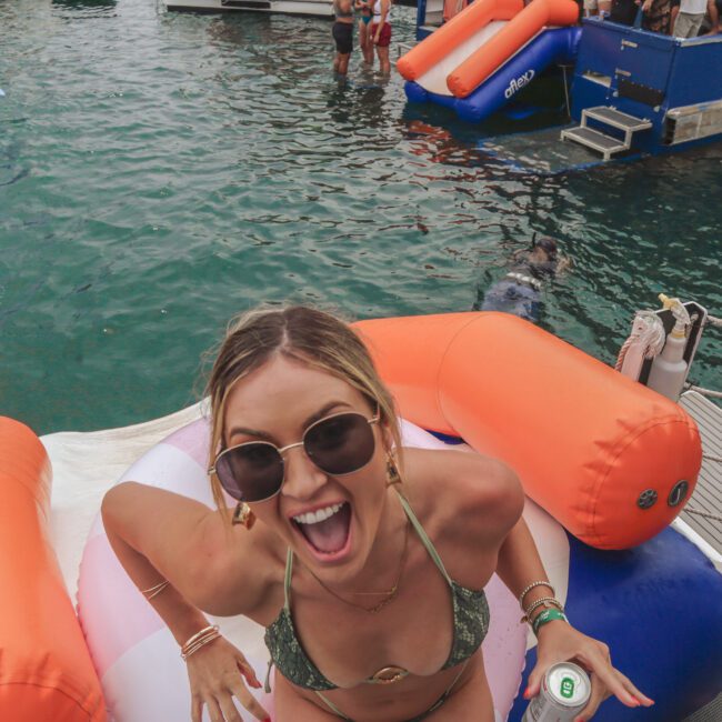 A smiling woman in sunglasses and a green bikini holds a drink while sitting on an inflatable float in the water near a docked boat. Other people swim and socialize nearby, with inflatables and a blue boat in the background.