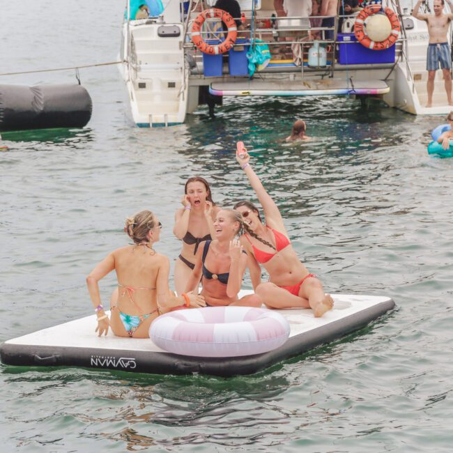 Four women in swimsuits sit and laugh on an inflatable raft in the water near boats. Other people relax and swim nearby in a festive, social atmosphere.
