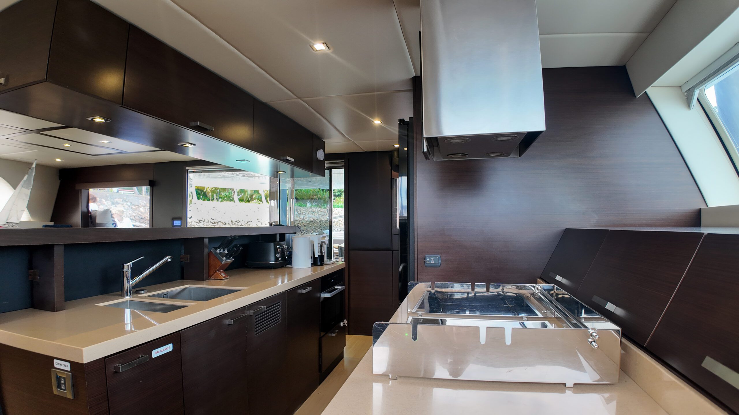Modern yacht kitchen with dark wood cabinets, beige countertops, stainless steel sink, stove, appliances, and large window letting in natural light. The kitchen appears tidy and well-organized.
