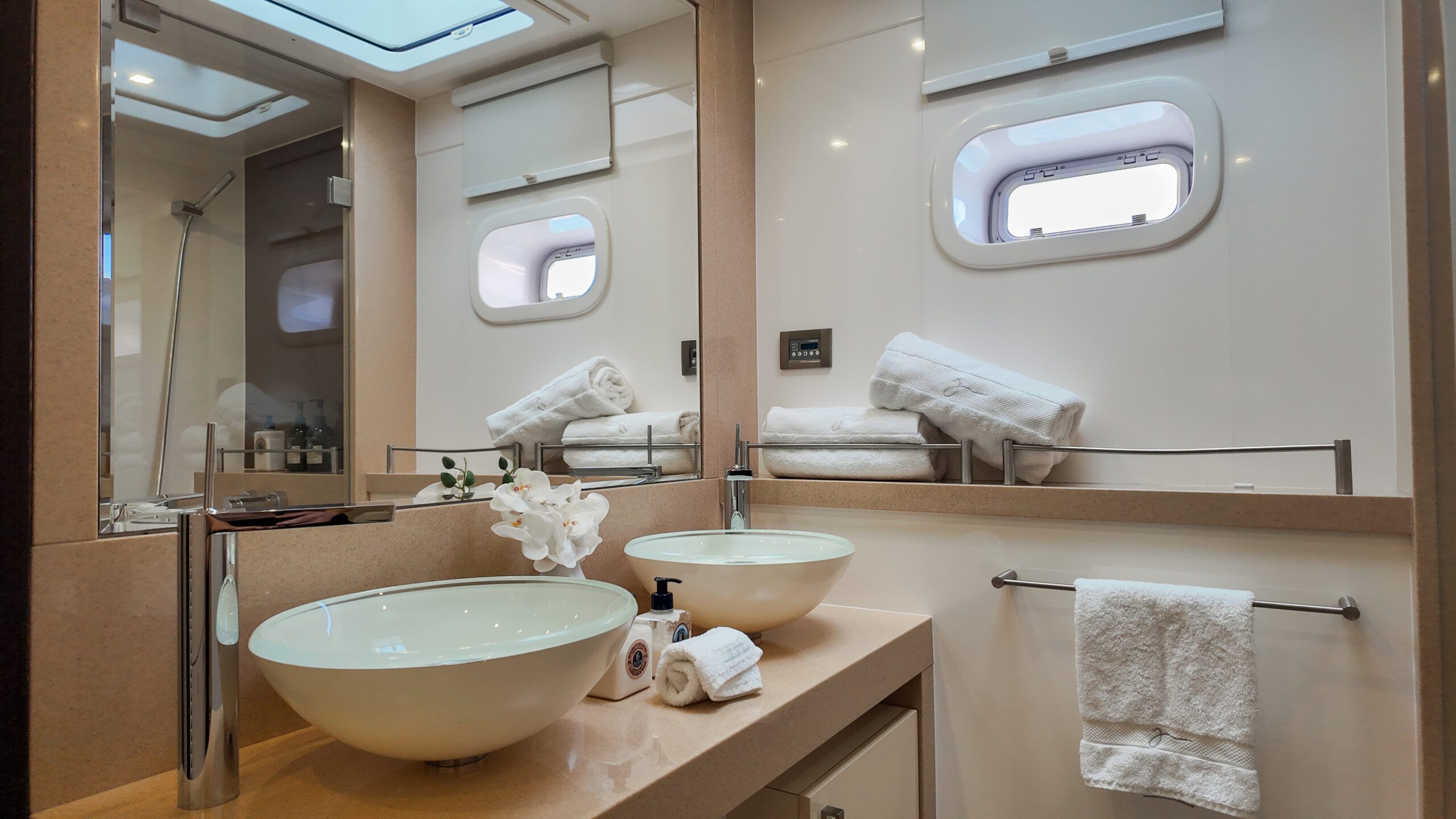 Modern bathroom with two round vessel sinks, a large mirror, stacked white towels, a window, and a rolled towel and orchid flower on the counter. The space is well-lit and has a clean, minimalist design.