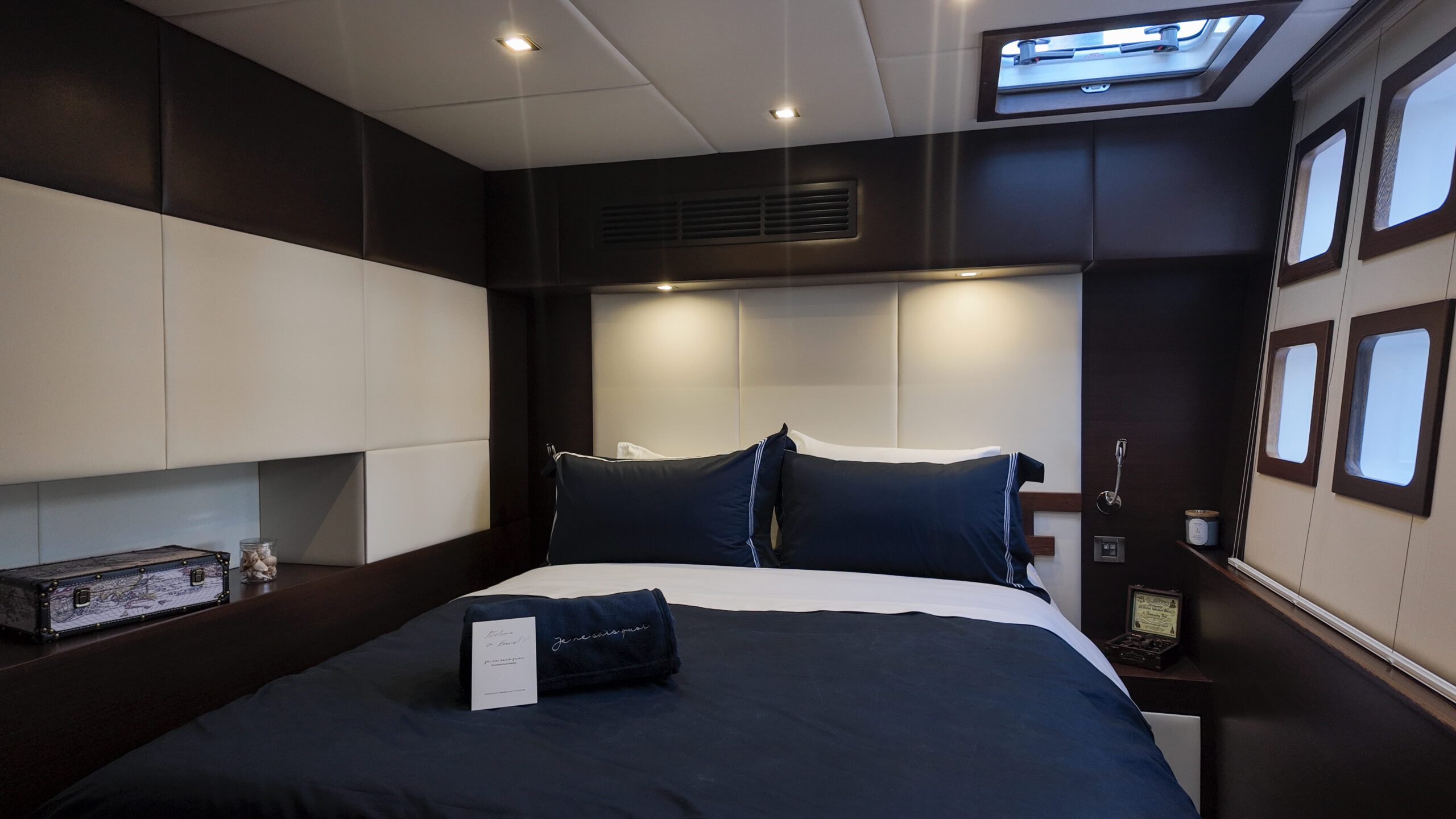A modern bedroom with a neatly made bed, navy blue bedding, and two pillows. There is a skylight above, several windows to the right, and a small table with a lamp and a book on the left side of the bed.