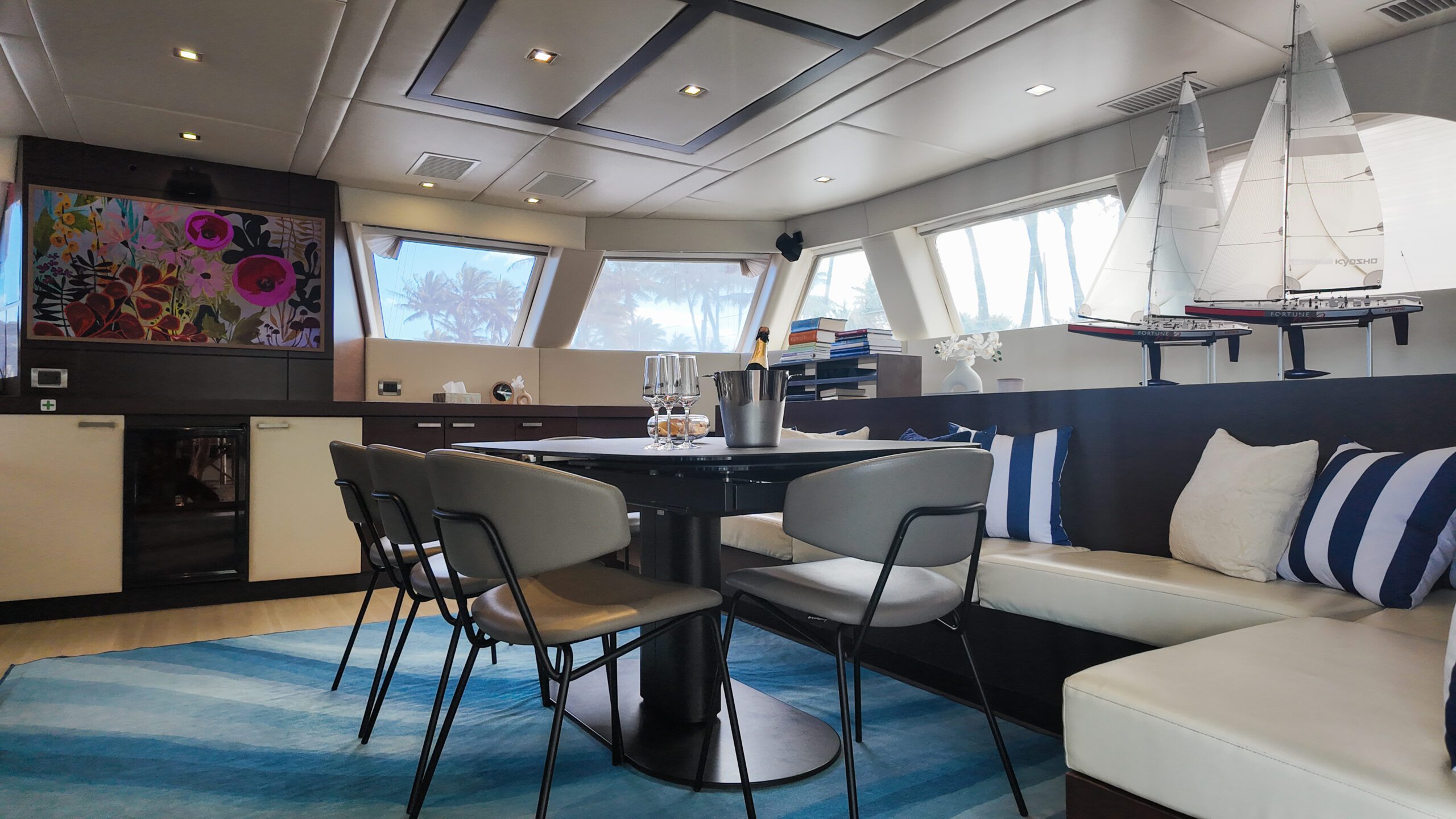 Modern yacht interior featuring a dining table with chairs, a curved bench with striped pillows, model sailboats, colorful wall art, and large windows with a view of palm trees outside.