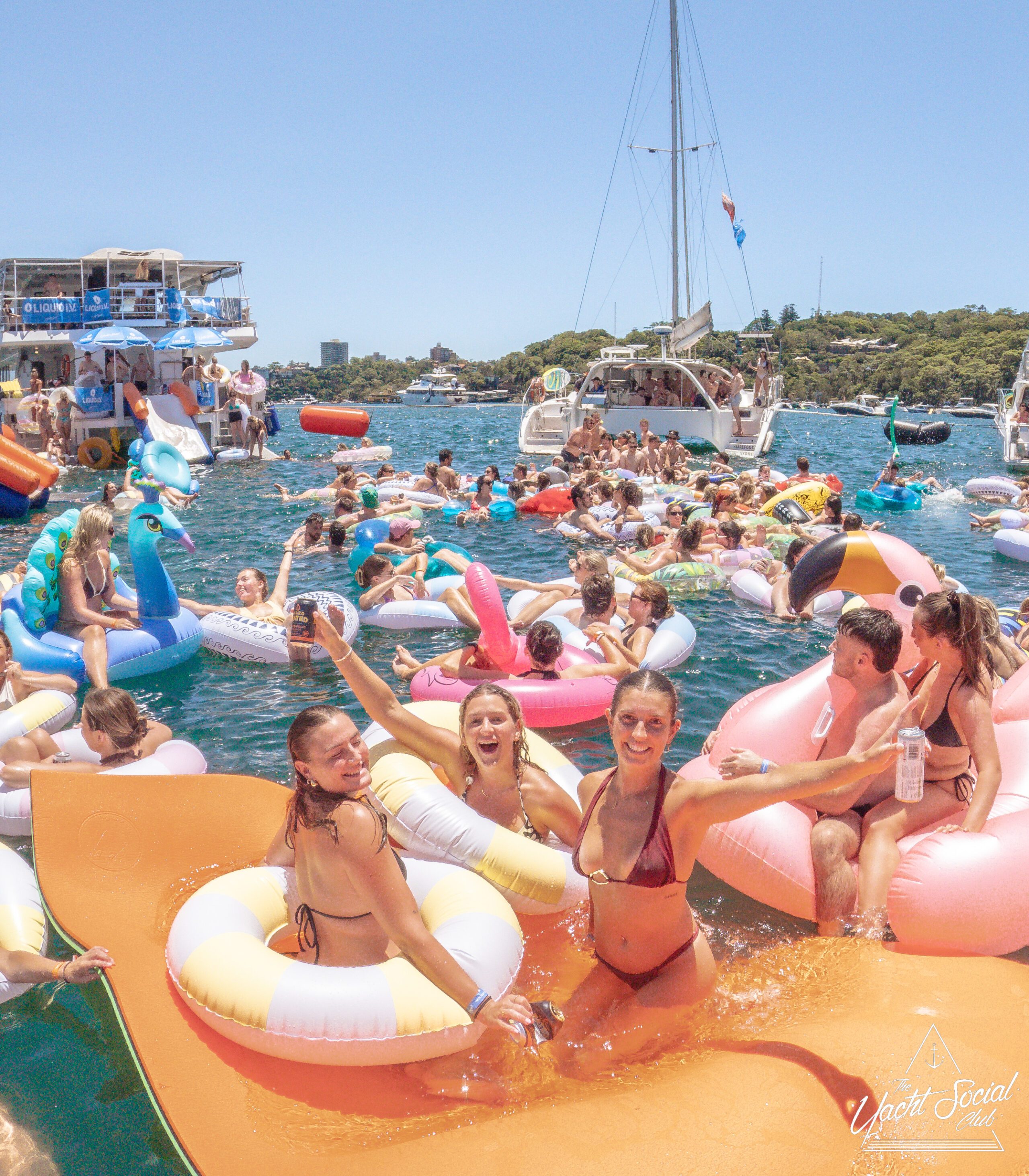 A lively group of people in swimsuits float on colorful inflatables at a sunny yacht party, with boats and clear blue water in the background. Everyone is smiling and enjoying the festive summer atmosphere.