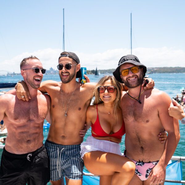 Four friends in swimwear stand arm-in-arm, smiling on the DJ Boat Male with water, yachts, and blue sky behind them. The relaxed group enjoys a sunny party or gathering on the water.